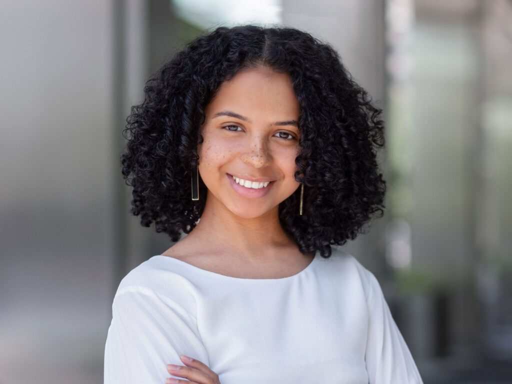 A young woman with curly black hair and a radiant smile stands confidently with her arms crossed. She is wearing a white top and is set against a blurred modern background, embodying a sense of cycle harmony.