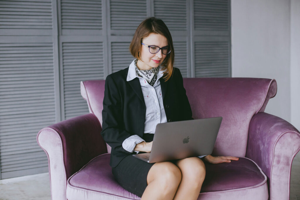 A woman in a business suit sitting on a plush purple sofa, working on a laptop. She has short hair and wears glasses, with a stylish scarf around her neck, focused on her screen in a contemporary workspace, embodying a sense of cycle harmony.